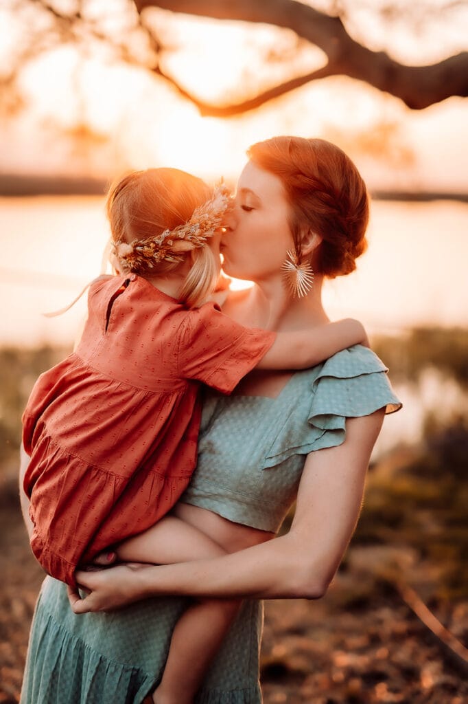 Family Photographer, a mother holds her baby girl close to her near a lake at sunset